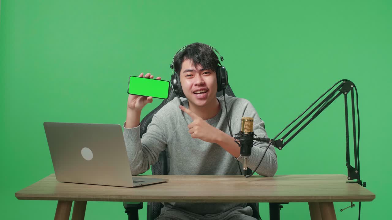 Smiling Asian Man With Headphone And Computer Showing Green Screen Mobile Phone While Sitting In Front Of Green Screen Background
