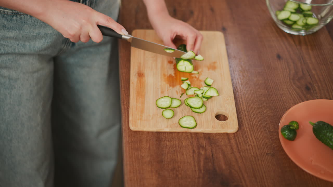 Overhead view of white girl slicing cucumber on wooden cutting board while preparing ingredients, with glass bowl containing cucumber slices and whole cucumber resting on orange plate