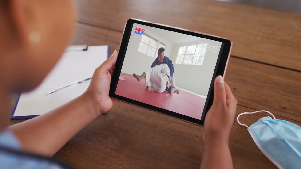 compuesto de una mujer sentada en una mesa con una máscara facial, viendo un partido de judo en la tableta