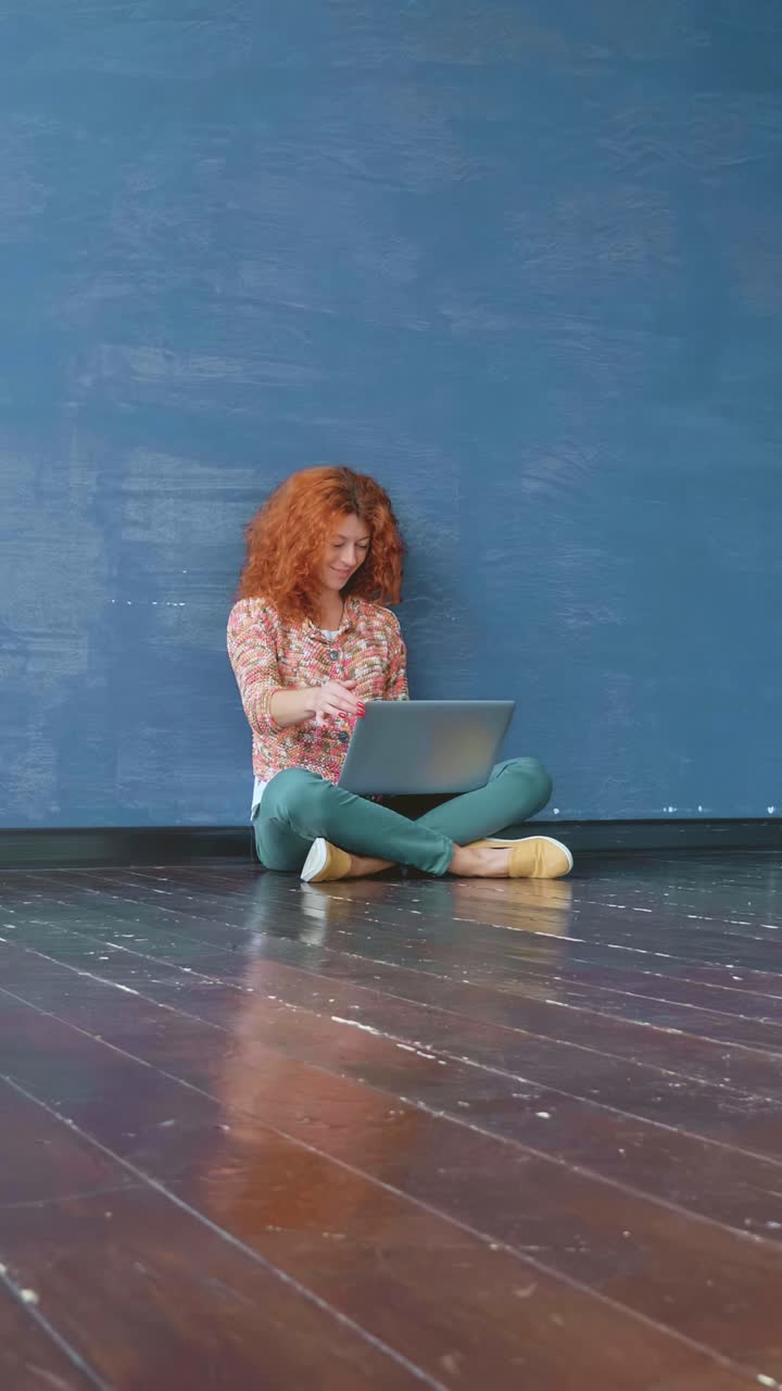 Young woman with curly red hair is sitting on wooden floor, working on laptop against a blue wall, showcasing a relaxed and creative workspace atmosphere