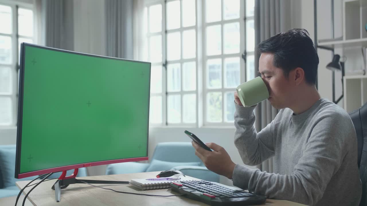 Asian Man In Long Sleeved T-Shirt With Green Screen Desktop Computer Using Mobile Phone And Drinking Coffee While Working At Home.