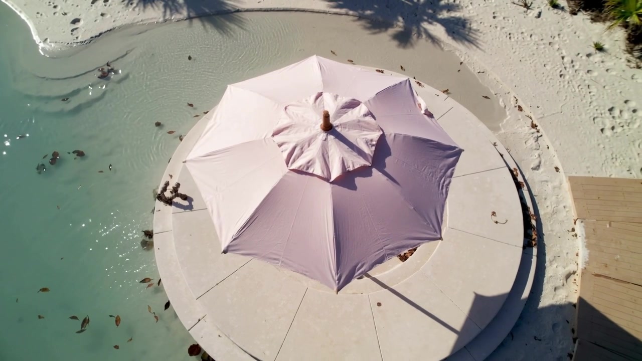 Drone che sorvola e filma un ombrellone rosa su una piattaforma rotonda a riva, con adulti in costume da bagno che guadano per rinfrescarsi.