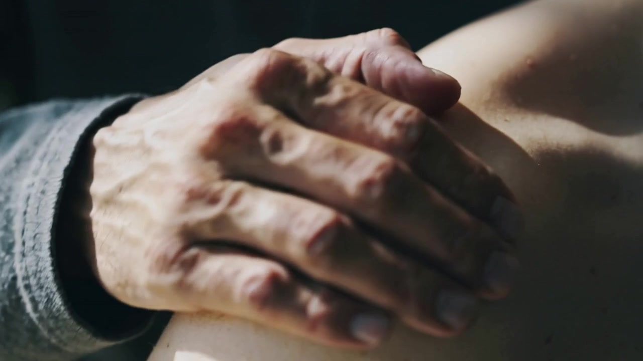 Entrata a sinistra in una stanza in penombra, mano destra che preme e accarezza una spalla nuda, polsino grigio, gesto confortante.