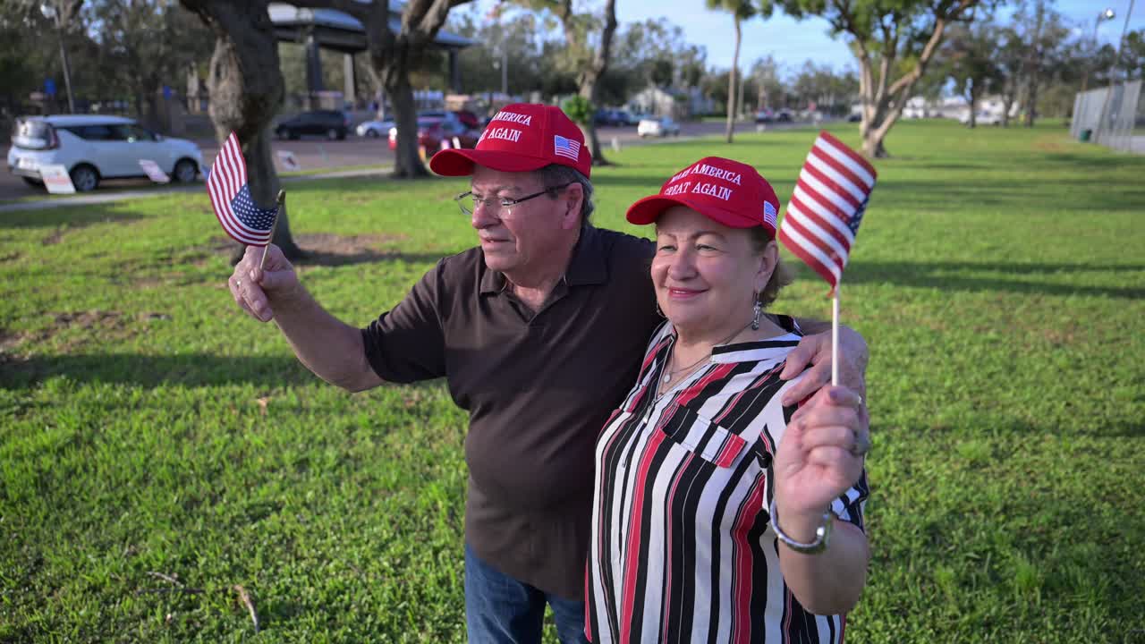 senior citizens wearing red MAGA hat wave flags at passing by traffic