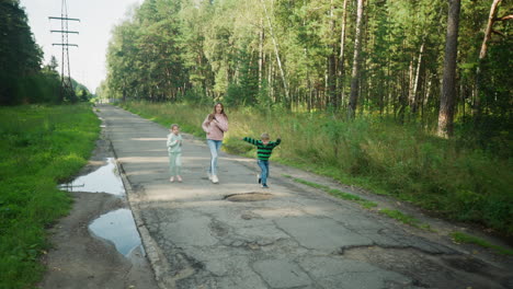 joyful young boy leaps over potholes on cracked countryside road while sister and older girl follow closely behind during fun outdoor moment surrounded by trees, greenery, and bright natural daylight