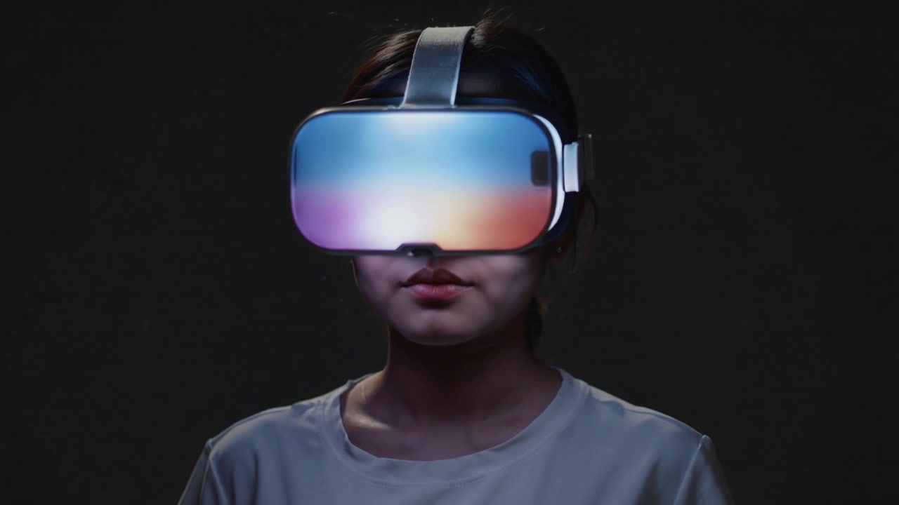 Ragazza adolescente che reagisce, con la testa leggermente inclinata, mentre il visore VR cambia colore in uno studio buio, con una maglietta chiara.
