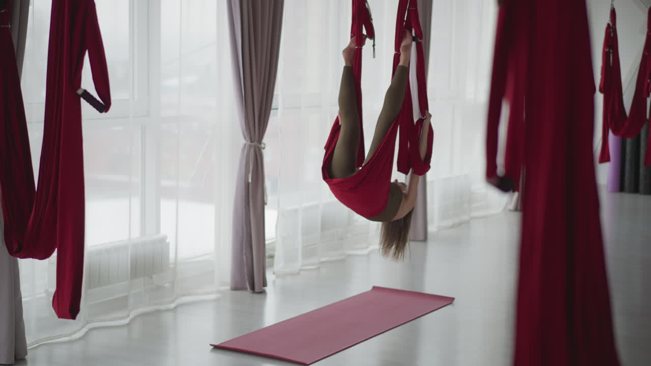 Flexible performer hanging inverted in red aerial silk hammock by floor to ceiling window curtains in bright modern studio, stretching body to show core strength balance flow and serene mindfulness