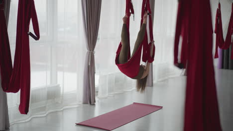 flexible performer hanging inverted in red aerial silk hammock by floor to ceiling window curtains in bright modern studio, stretching body to show core strength balance flow and serene mindfulness