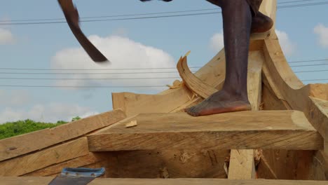 barefoot black man carving and cutting wooden boat with adze in ghana