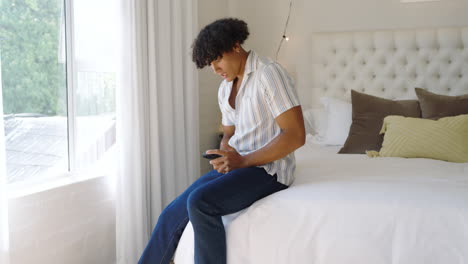 young man sitting on bed using smartphone, relaxing in bright bedroom, at home