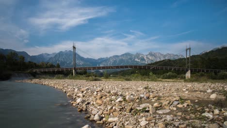 Lapso-De-Tiempo-Del-Paisaje-De-Los-Alpes-Italianos-Con-Un-Río-Que-Fluye,-Un-Puente,-Montañas-En-El-Fondo-Y-Clounds-Moviéndose