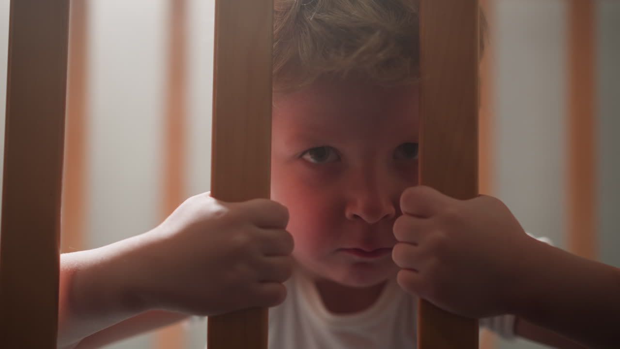 겁에 질린 작은 소년은 손으로 침대의 나무 막대기를 잡습니다.