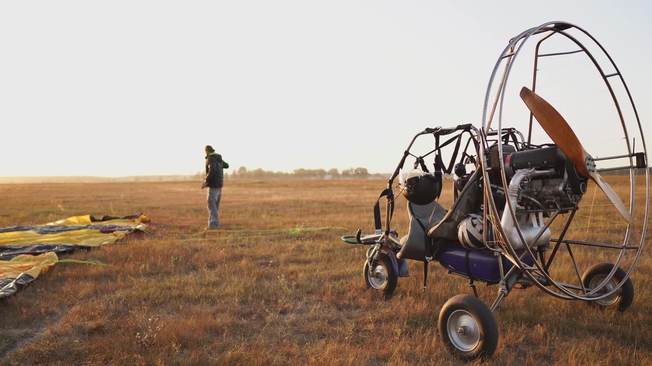 모터 패러글라이더는 해가 지는 시점에서 나무 프로펠러와 함께 필드에 서 있으며, 조종사는 낙하산을 놓고 슬링을 정렬합니다.