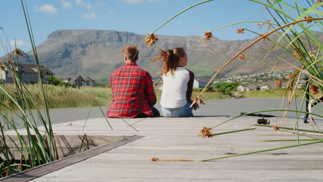 Couple-sitting-on-a-jetty-by-a-lake-and-mountains,-back-view