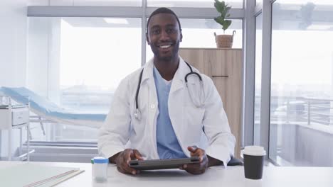 African-american-male-doctor-using-tablet-and-making-video-call-waving-in-hospital,-slow-motion