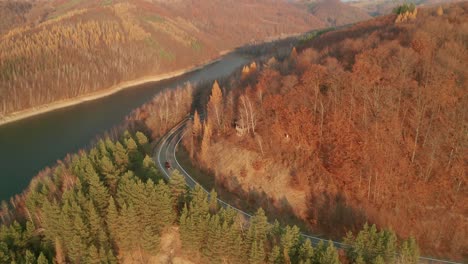 Eine-Absteigende-Luftdrohnenansicht-Eines-Autos,-Das-Im-Spätherbst-Auf-Einer-Kurvenreichen-Straße-Entlang-Eines-Sees-Fährt