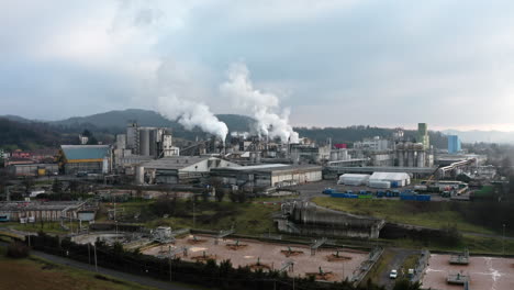 A large industrial factory emitting smoke, with pipelines, storage tanks, and surrounding hills