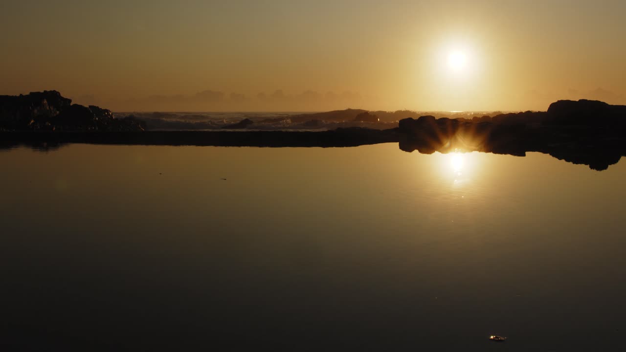 reflection of golden sunrise in coastal rock pool, surf breaking in background