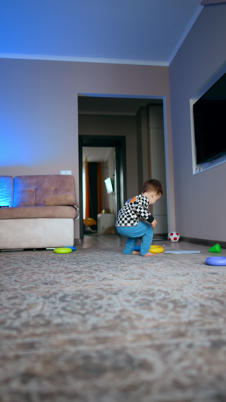 Baby boy playing in the room throws his toys around. Kid sits on the floor and rolls the plastic ring by the floor. Low angle view. Vertical video.