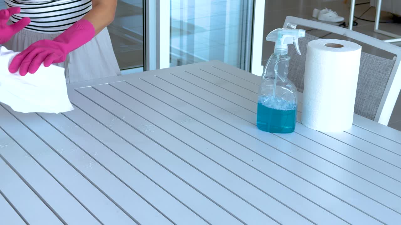 Person wearing pink gloves cleans a table using a spray bottle and paper towels in a bright room