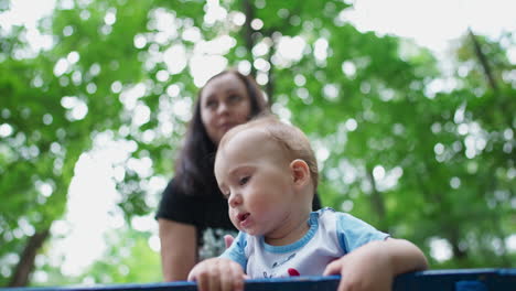 healthy caucasian child spending time outdoors. cute kid standing on the bench looking curiously around. close up. mom at backdrop. low angle view.