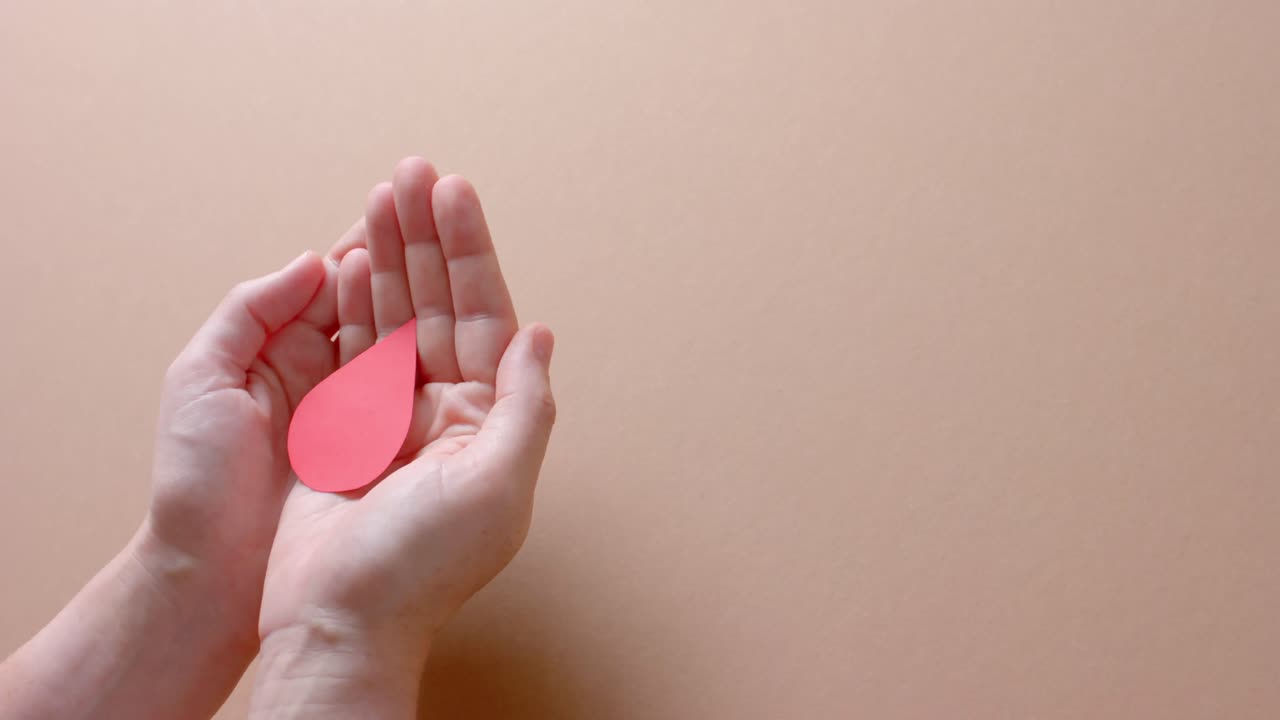 Hands of caucasian woman cupping blood drop on beige background with copy space, slow motion