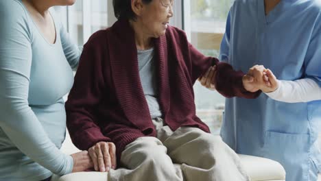 Asian-female-physiotherapist-treating-arm-of-senior-female-patient,-with-adult-daughter-at-surgery