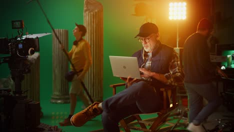 prominent successful director sitting in a chair on a break using laptop computer. on the studio film set with high-end equipment professional crew shooting high budget movie