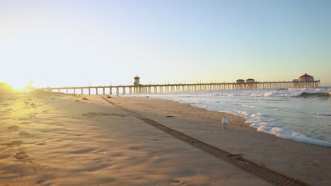 Ein-4K-Zeitraffer-Bei-Sonnenaufgang,-Wobei-Sich-Der-Pier-Im-Hintergrund-Bei-Sonnenaufgang-Von-Links-Nach-Rechts-Bewegt,-Während-Familien-Ihren-Urlaub-Genießen-Und-Surfer-Wellen-Fangen