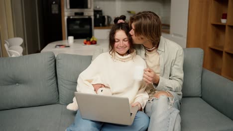 Young-adult-couple-talking-using-laptop-computer-at-home-sitting-on-couch,-watching-video