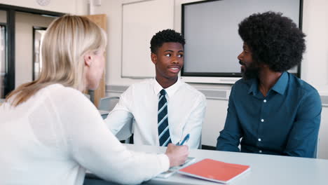 father and teenage son having discussion with female teacher at high school parents evening