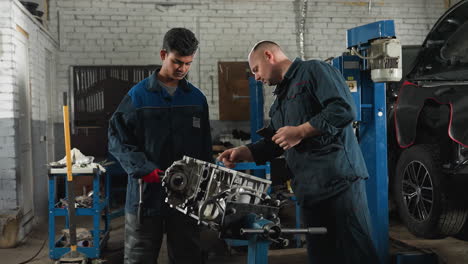 two mechanics in an automotive workshop; one consulting his phone to show something to his colleague who is focused on the engine block in front, surrounded by mechanical equipment