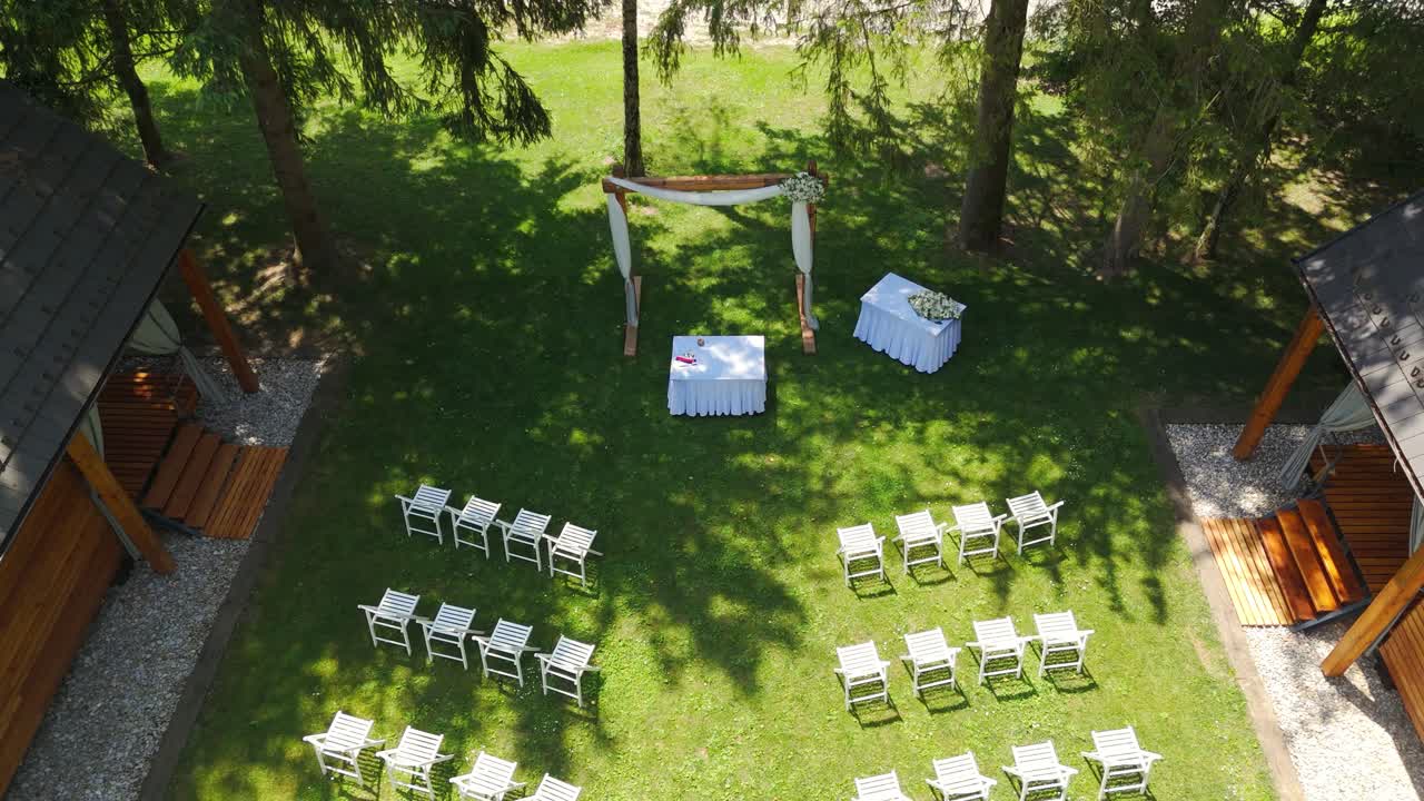 Aerial: wedding ceremony with an arch and white chairs during the day in the forest, crane down drone shot