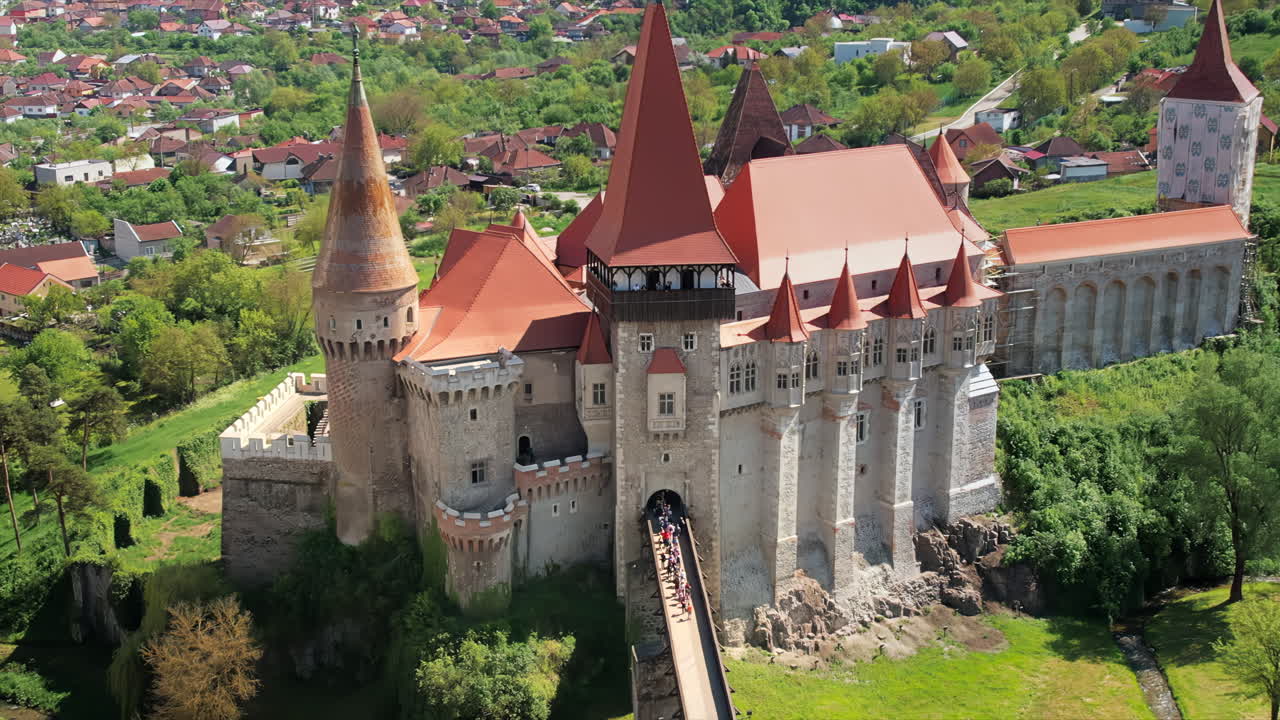 Аэрофотосъемка замка Корвин, расположенного в Хунедоаре, Румыния. Люди идут по мосту, ведущему ко входу. Вокруг — зелень и здания.
