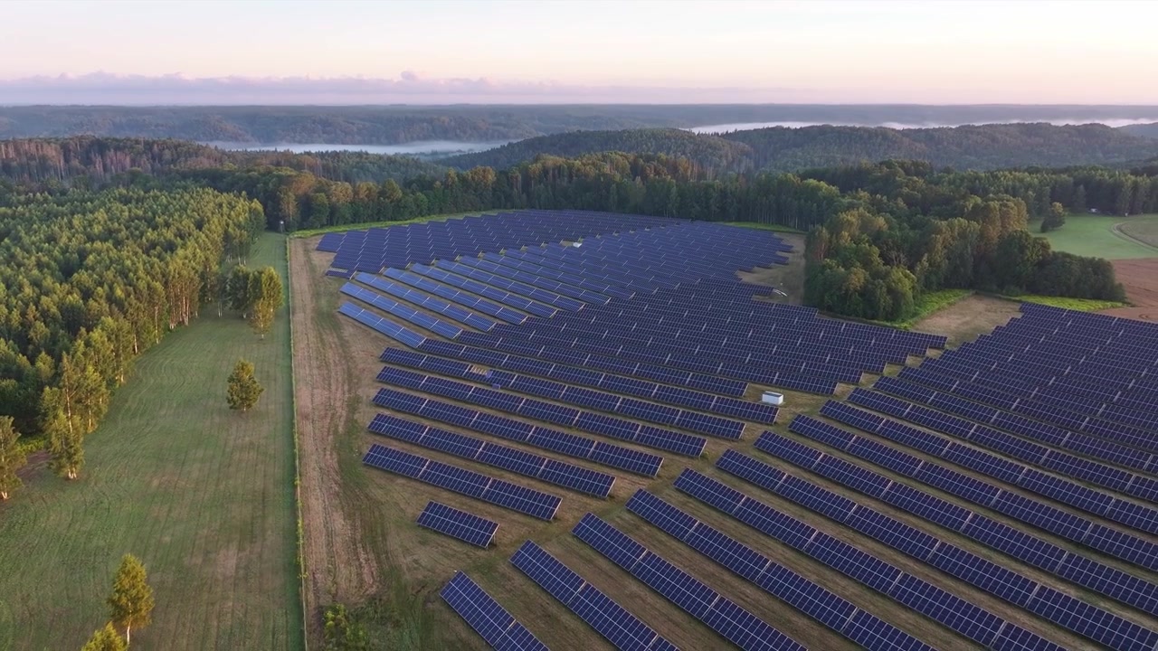 Droneoptagelser fra luften af en stor solcellepark nær Sigulda, Letland, omgivet af skove og landbrugsjord, med søer og morgentåge synlig i det fjerne