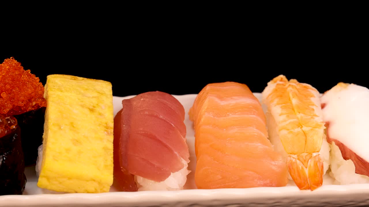 A close-up shot of a sushi platter with various nigiri and sashimi pieces slowly rotating against a black background under bright, even lighting