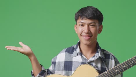 close up of asian teenager with guitar smiling and pointing to the side on green screen background