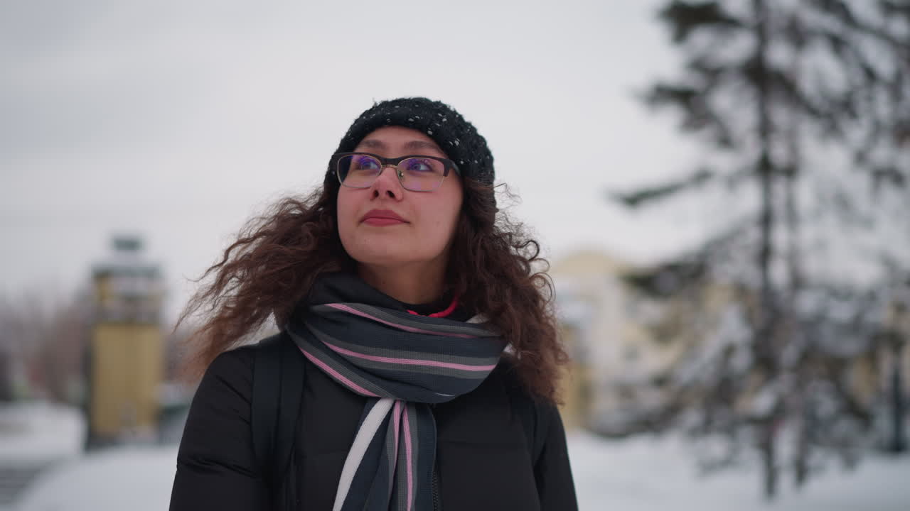Female wearing glasses winter coat scarf and knitted hat standing outdoors in snowy urban street looking aside with thoughtful expression curly hair blowing in cold wind blurred buildings in background