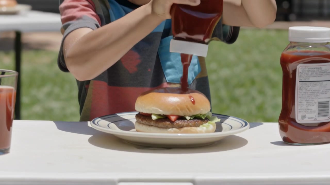 Bambino con maglietta a righe e bavaglino blu che spreme una bottiglia di ketchup, aggiungendolo a un hamburger in giardino.