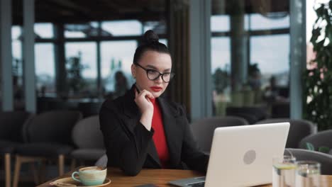 Mujer-Joven-Con-Gafas-Y-Aparatos-Ortopédicos,-Trabajadora-Independiente,-Sentada-En-Un-Elegante-Restaurante-Vestida-Con-Traje-De-Negocios,-Trabajando-Atenta-Y-Pensativamente-En-Su-Computadora-Portátil.