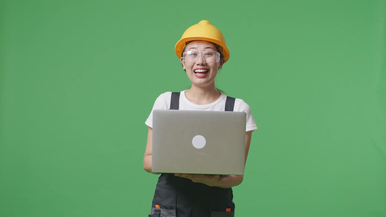 녹색 스크린 배경 스튜디오에 서 있는 아시아 여성 노동자가 노트북을 사용하고 미소 짓고 보호 안경과 안전 헬을 착용하고 있습니다.