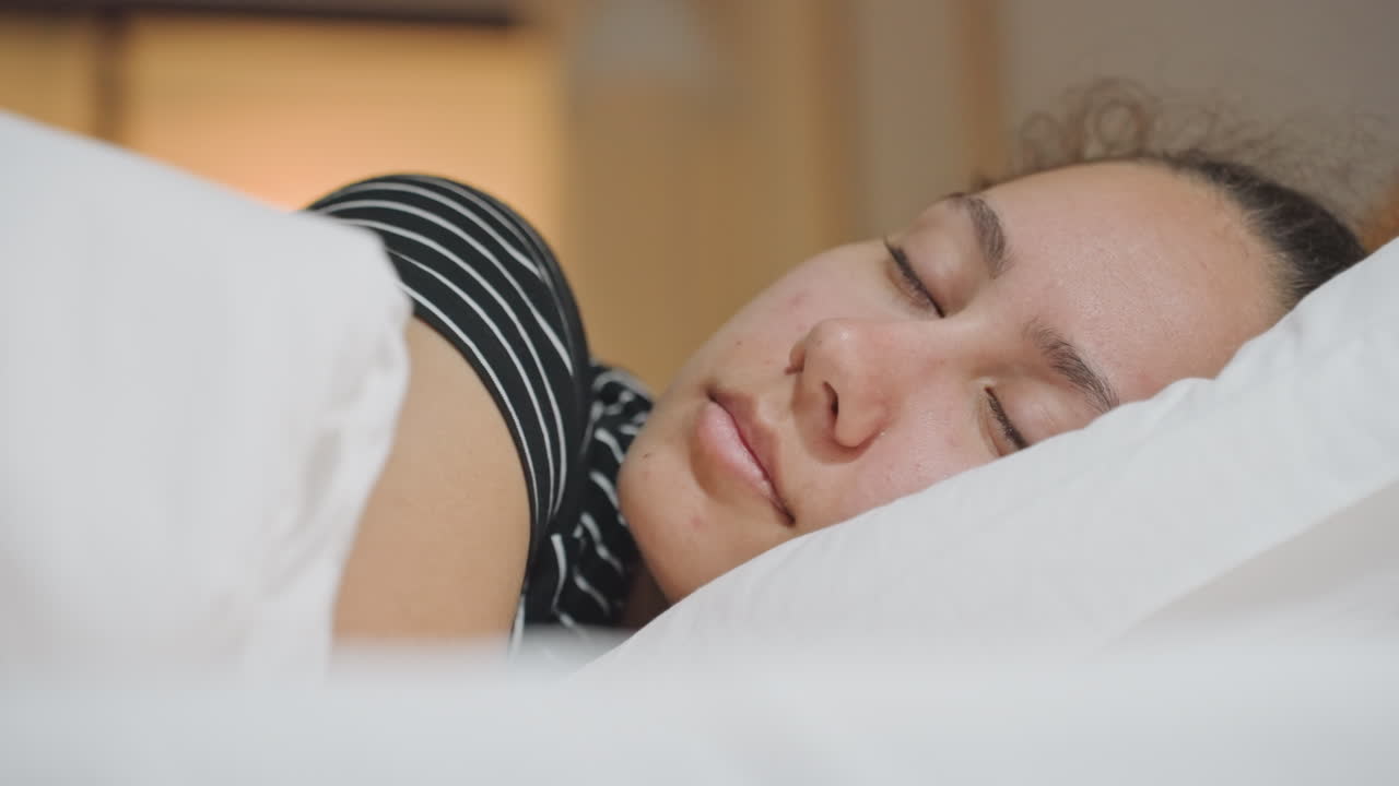 Blandingsrace forretningsrejsende sover på hotel, nærbillede, der viser hvid pude, stribet top, varmt lys fra sengebord og roligt udtryk – en intim gæstfrihedsscene, der indfanger hvile, afslapning og stille restitution.