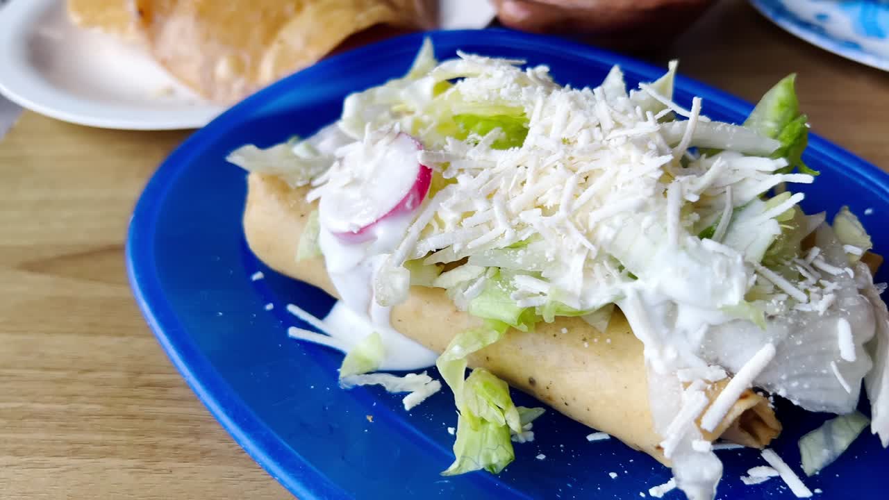 Delicious flautas, typical mexican food, lying on a blue plate and covered with shredded cheese, cream, lettuce and radish slices, ready to be eaten