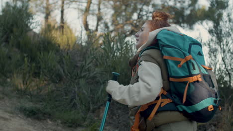 Side-View-Of-A-Red-Haired-Female-Backpacker-With-Trekking-Poles-Hiking-In-The-Forest-On-A-Sunny-Day