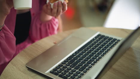 a person using a laptop on a wooden table takes a coffee cup in one hand while holding a burger in the other, they carefully place the coffee cup back on the table and resume working on the laptop