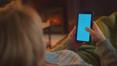 woman at home lying on sofa in lounge with cosy fire using blue screen mobile phone