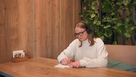 Woman wearing glasses and black headphones sits at wooden table writing on napkin while enjoying music and gently dancing, surrounded by cozy interior with wood panel wall