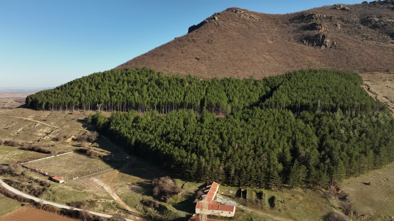 heart-shaped pine forest at the base of a rocky hill, with rural buildings nearby. Greece