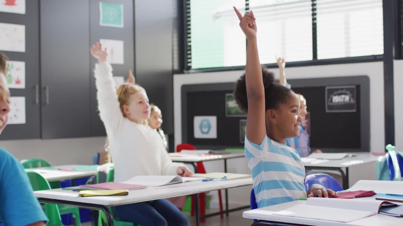 Diverse happy schoolchildren sitting at desk and raising hands in school classroom