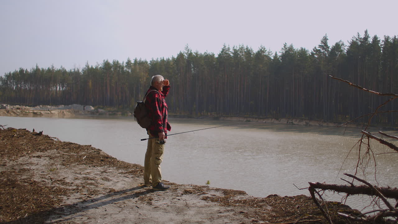 aged fisher is viewing reservoir in forest standing on high coast at sunny autumn day angling and spin fishing
